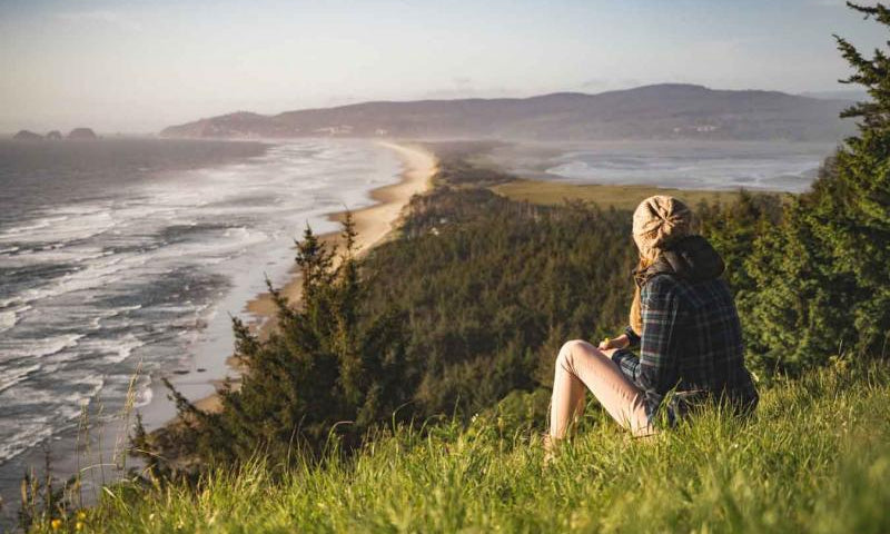 Woman sitting on a hill overlooking a tree-filled ocean shoreline.